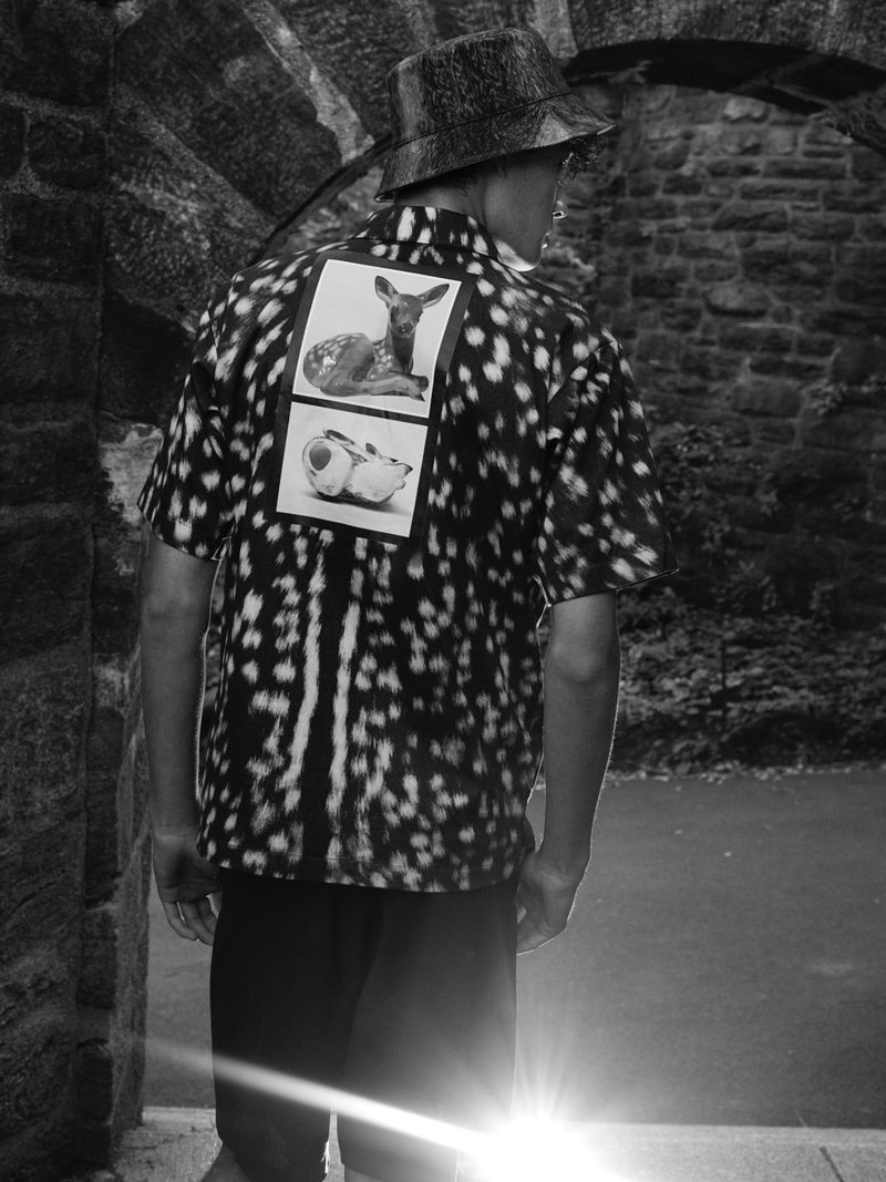 Back view of a person wearing a patterned shirt and hat, standing in a stone archway with bright sunlight, black and white photograph.