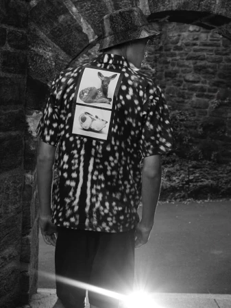 Back view of a person wearing a patterned shirt and hat, standing in a stone archway with bright sunlight, black and white photograph.