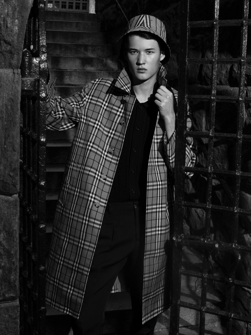 Young adult poses in a plaid trench coat and bucket hat by a stone staircase behind an iron gate in a black and white fashion shot.
