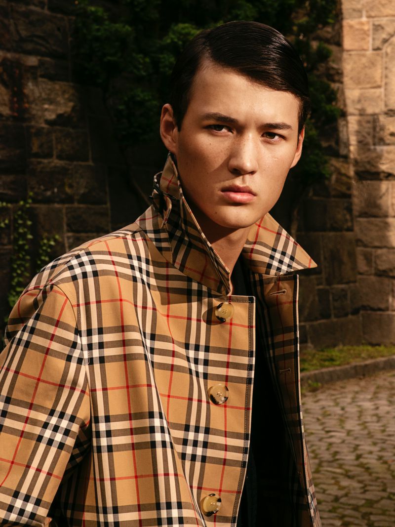 Male model poses in a Burberry trench coat against a stone wall backdrop. The shot captures a fashion moment in natural lighting.