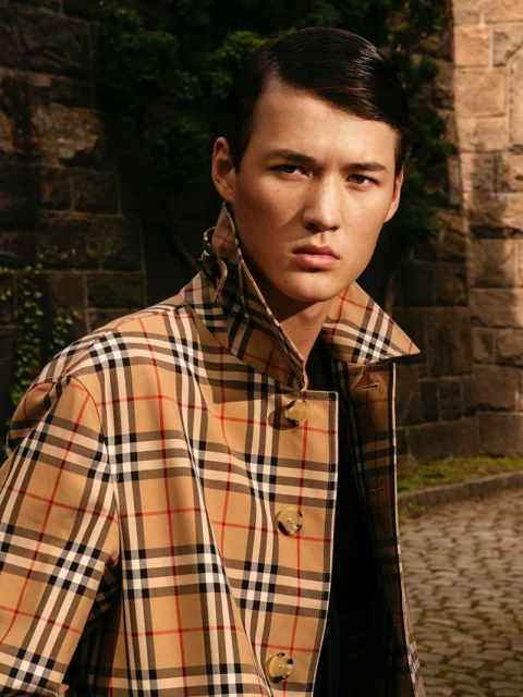 Male model poses in a Burberry trench coat against a stone wall backdrop. The shot captures a fashion moment in natural lighting.