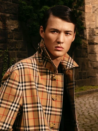 Male model poses in a Burberry trench coat against a stone wall backdrop. The shot captures a fashion moment in natural lighting.