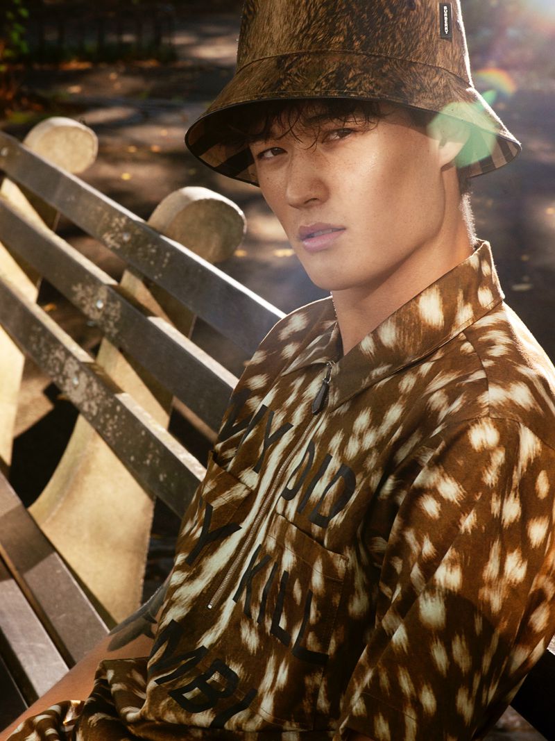 Close-up portrait of a young adult male wearing a patterned shirt and bucket hat, sitting on a wooden bench in an outdoor setting.
