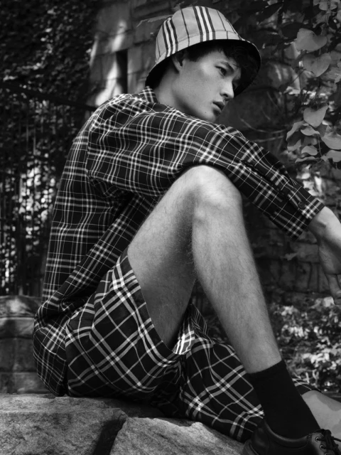 Monochrome shot of Asian male model in plaid bucket hat, shirt, and shorts, sitting on stone steps in an outdoor setting with foliage.