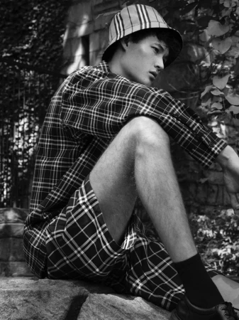 Monochrome shot of Asian male model in plaid bucket hat, shirt, and shorts, sitting on stone steps in an outdoor setting with foliage.
