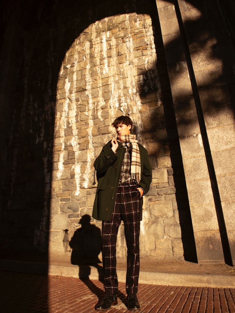 A young man stands in front of a brick wall wearing a green coat, plaid pants, and a scarf in an urban setting with dappled sunlight.