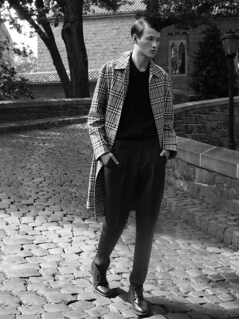 Male fashion model in plaid trench coat and boots posing on cobblestone street in black and white, with stone buildings and trees.