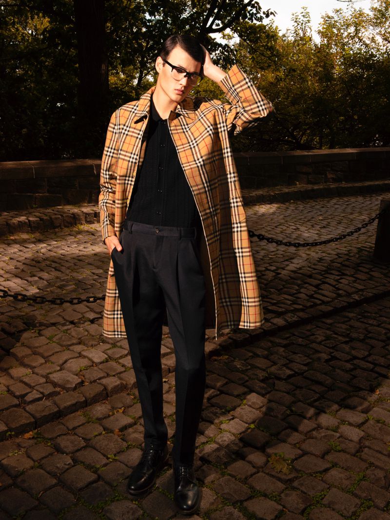 Male fashion model in plaid coat, black shirt, and dark pants poses on cobblestone path surrounded by trees. Autumn day, outdoor setting.