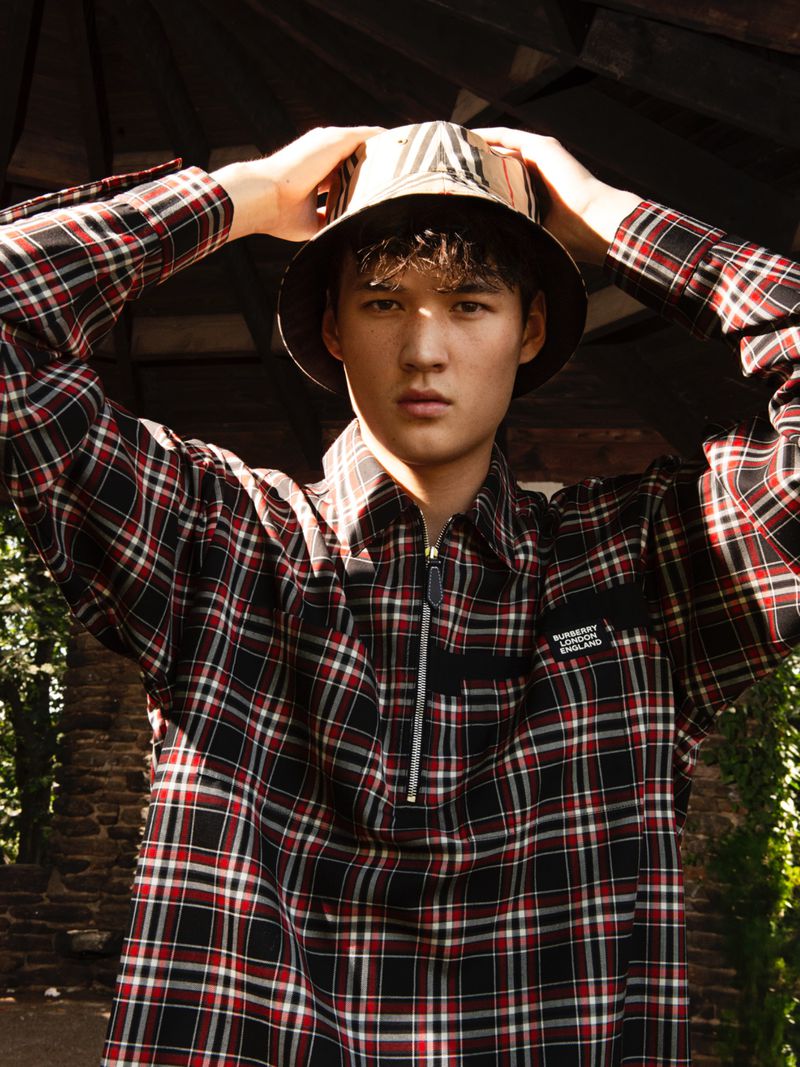 Male model poses in a plaid shirt and Burberry bucket hat in an outdoor setting with trees and shadows, showcasing fashion.