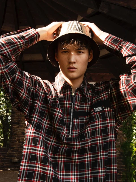Male model poses in a plaid shirt and Burberry bucket hat in an outdoor setting with trees and shadows, showcasing fashion.