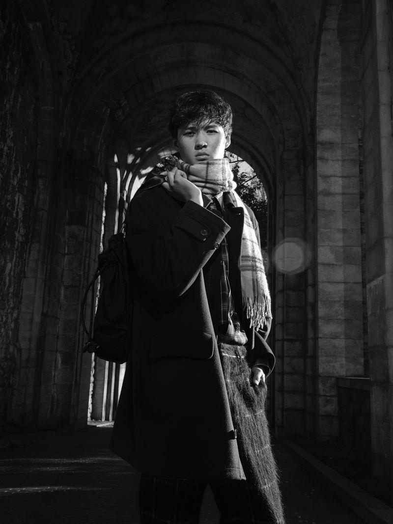 Monochrome portrait of a young man in a coat and scarf standing in an arched stone hallway with light at the end of the tunnel.
