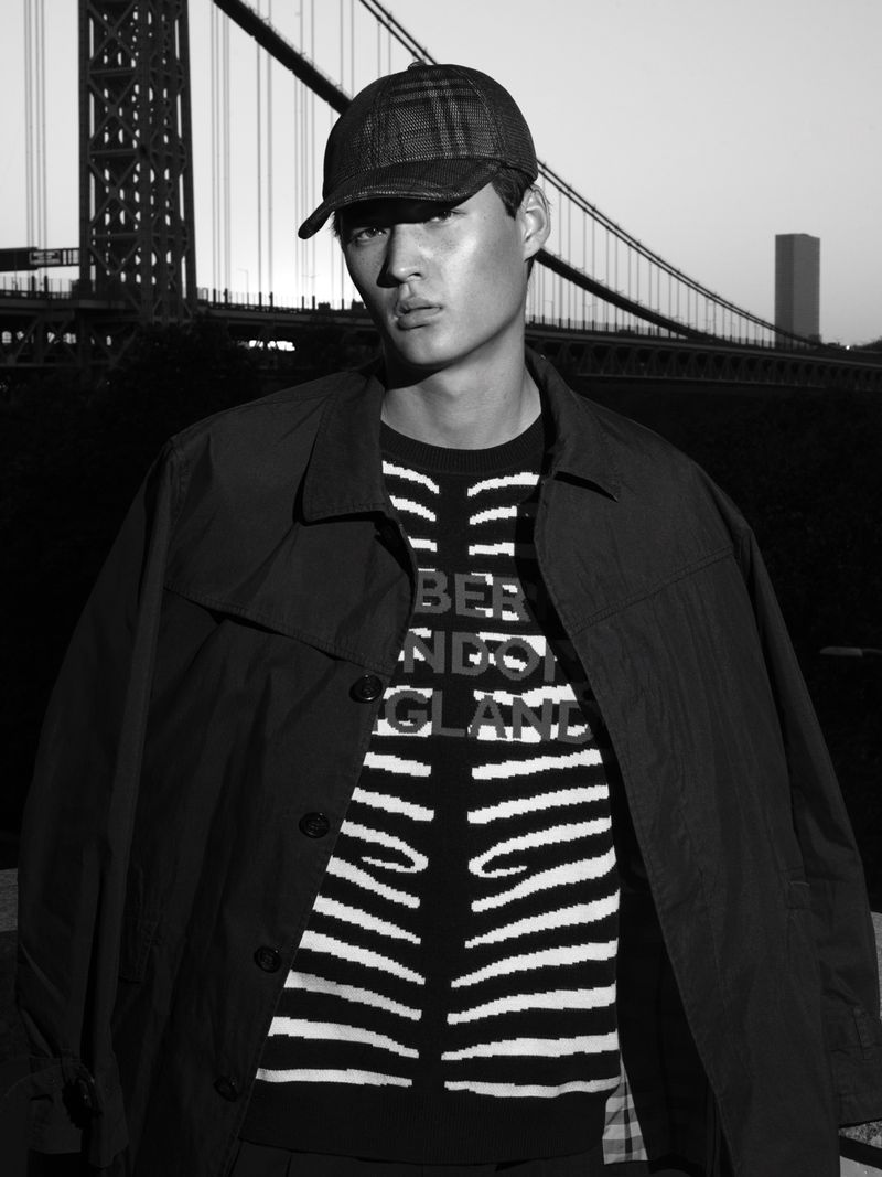 Portrait of a young adult male model wearing a patterned cap, jacket, and sweater, posing against a blurred urban bridge background.
