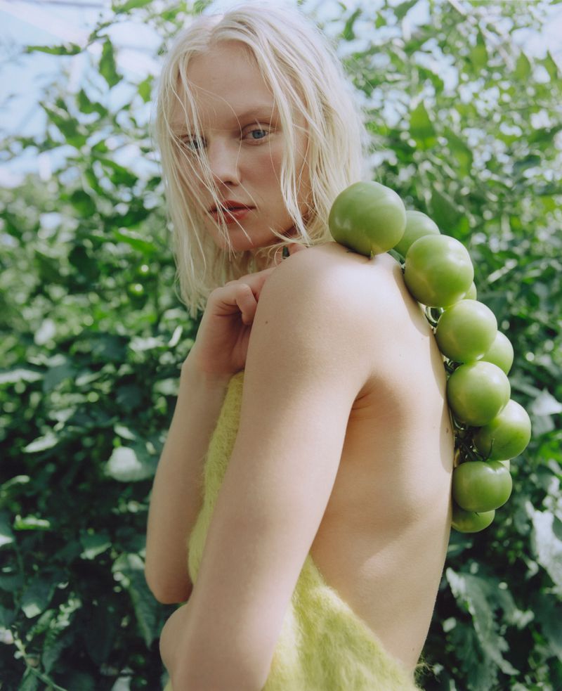 Blonde female model poses with a string of green tomatoes in an outdoor setting, wearing a textured, yellow garment.