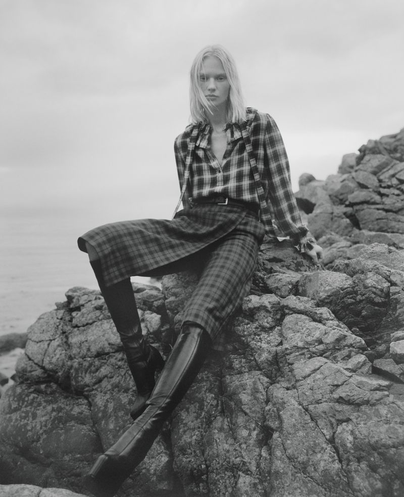 Monochrome fashion portrait of a person with blonde hair in a plaid top and skirt sitting on rocks near the ocean under an overcast sky.