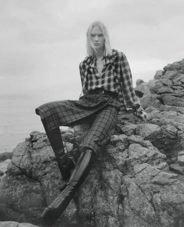 Monochrome fashion portrait of a person with blonde hair in a plaid top and skirt sitting on rocks near the ocean under an overcast sky.