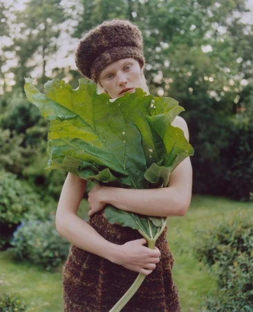 Model Vilma Sjöberg wears a textured brown knit hat and skirt while holding a large green leaf in a lush outdoor garden setting.