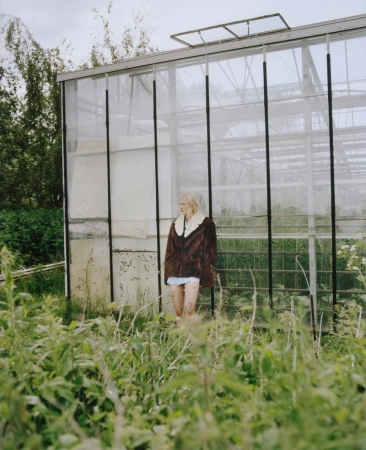 Young woman in a brown coat standing in a lush green field in front of a greenhouse on an overcast day, creating a serene outdoor portrait.