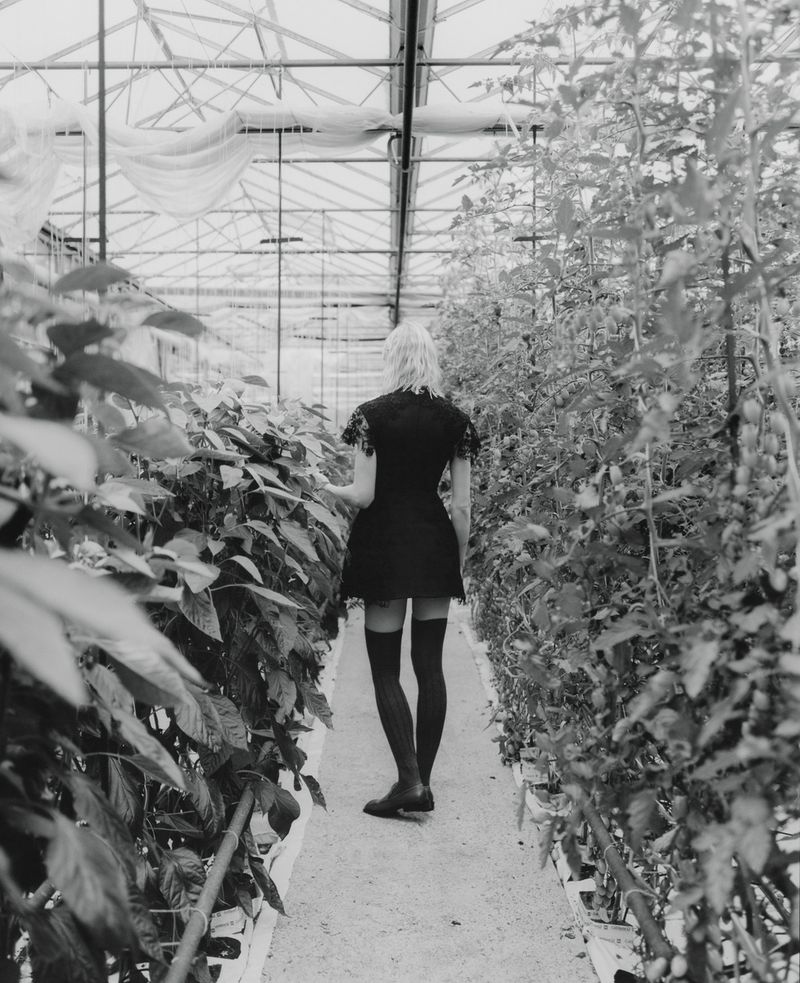 Vilma Sjöberg seen from behind walking down a greenhouse aisle wearing a black mini dress and knee-high socks in black and white.