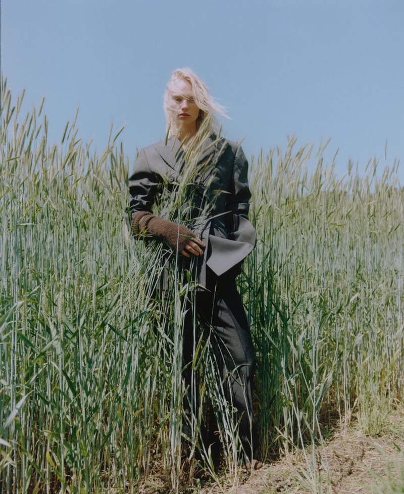 Fashion editorial shot of a fair-skinned blonde woman in a blazer and trousers standing in a wheat field under a blue sky on a sunny day.