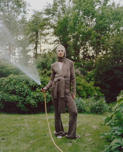 Woman in brown knitwear holding hose, watering lush green lawn and garden on overcast day. Fashion editorial outdoor shot. 