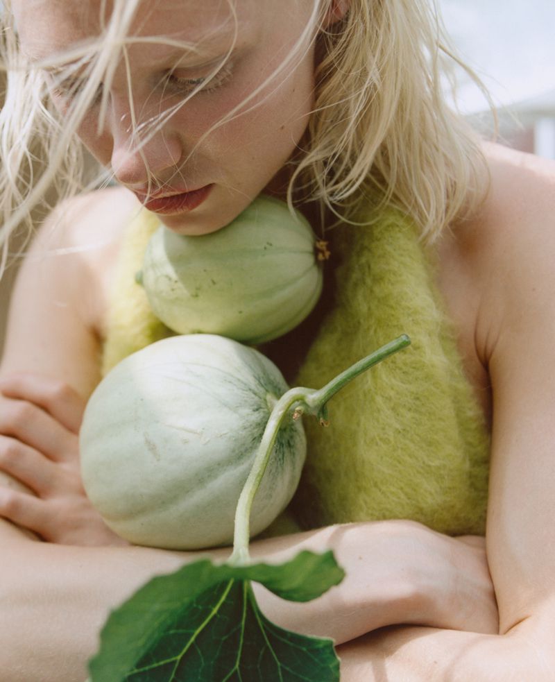 Blonde woman embraces two light green melons in a close-up shot, wearing a fuzzy yellow-green sweater, outdoors with natural light.