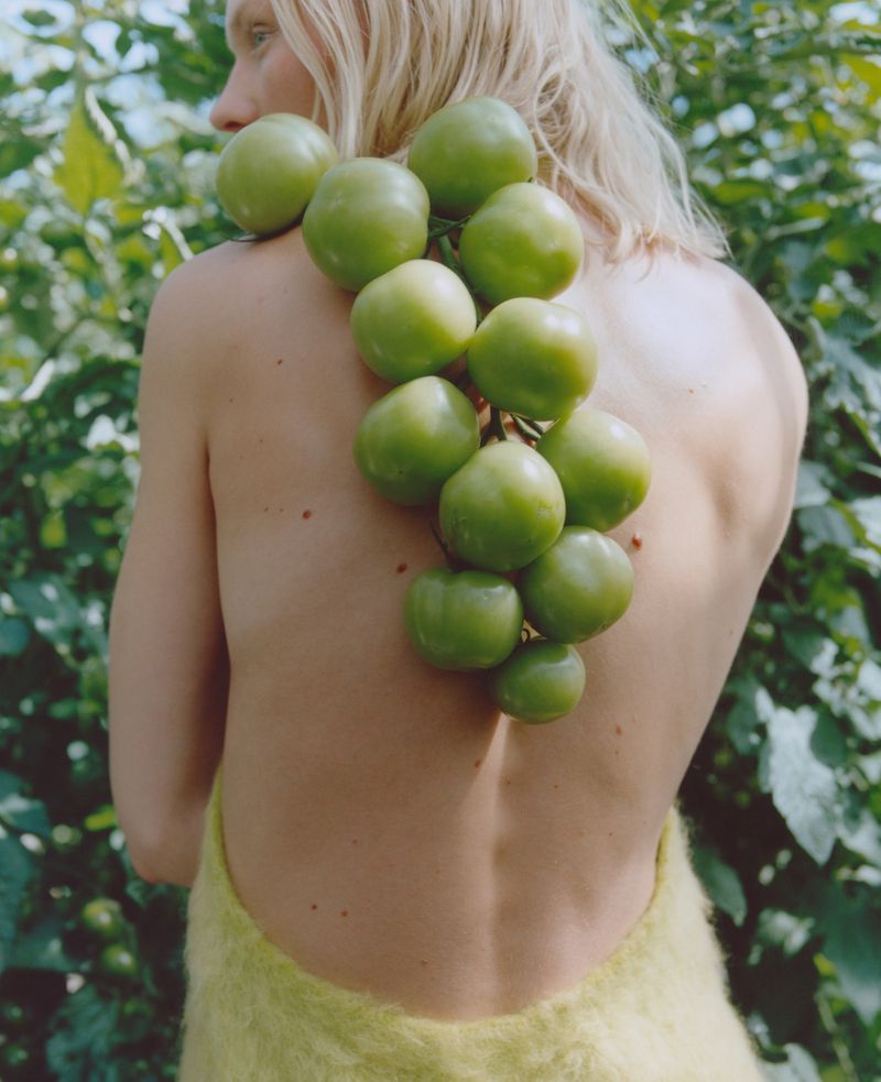 Blonde woman with green tomatoes on her back posing in a natural setting with foliage; close-up shot capturing the texture of tomatoes and leaves.