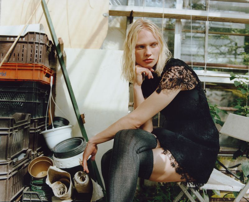 A blonde woman in a black lace dress and gray socks sits on a chair in a greenhouse with gardening crates and plants.