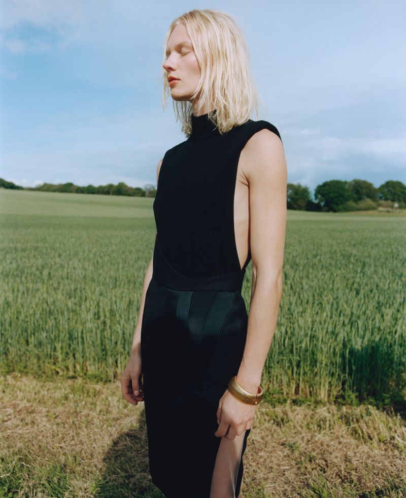 Model Vilma Sjöberg in a sleeveless black high-neck dress and gold cuff bracelet standing outdoors in an open green field.