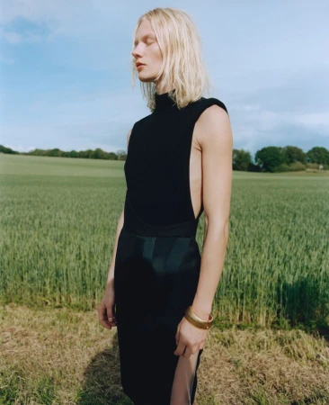 Model Vilma Sjöberg in a sleeveless black high-neck dress and gold cuff bracelet standing outdoors in an open green field.