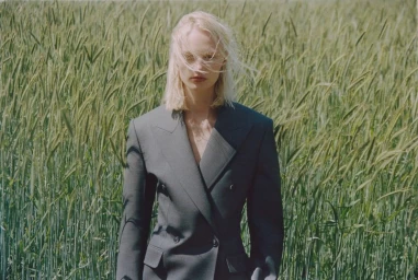 Model Vilma Sjöberg in a tailored charcoal blazer with windblown blonde hair standing in a tall green wheat field.