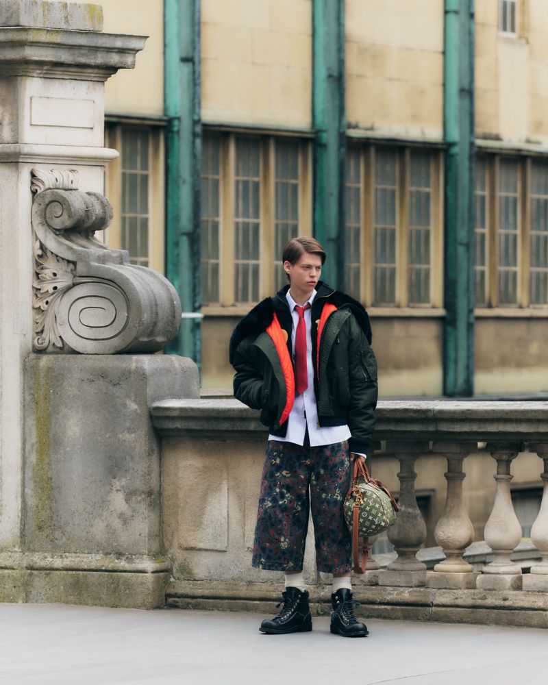 Young adult male fashion model poses outdoors wearing a green bomber jacket, white shirt, red tie, floral pants, and black boots.