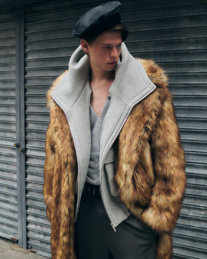 Male model in brown fur coat, grey sweater and black beret poses against a gray metal roll-up door, fashion photography, outdoor setting.