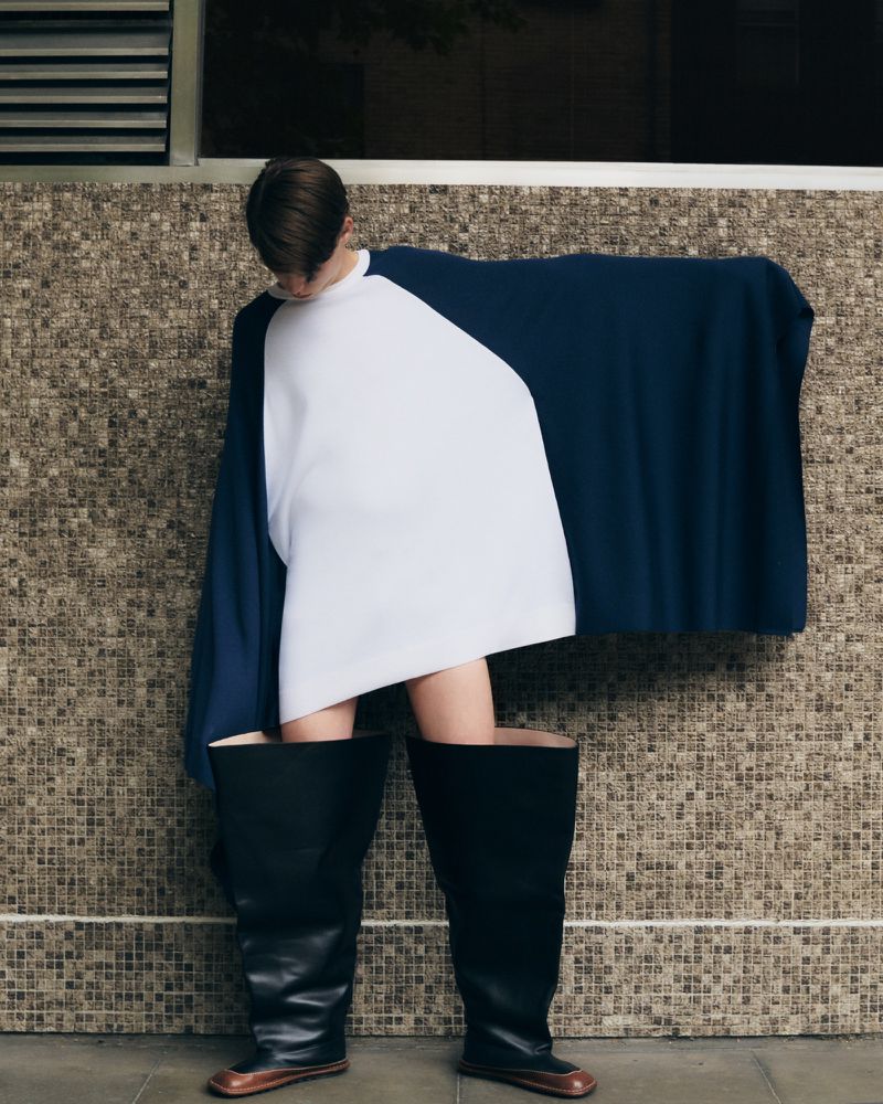 Person poses in an unusual outfit: a white and navy top paired with oversized black boots against a textured beige wall. Fashion shot.