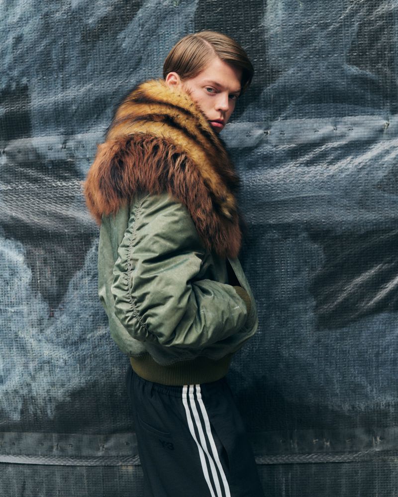 Male fashion model poses in an olive green bomber jacket with fur collar and black Adidas track pants against a textured gray wall.