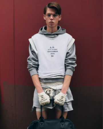 Male model poses in gray hoodie, white sleeveless top with black lettering, and gray shorts against a red wall, holding a black duffel bag.