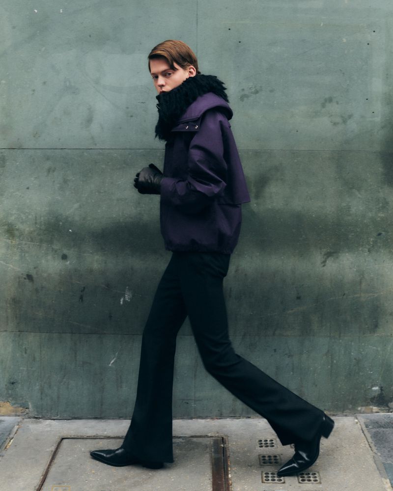 Stylish man in a purple jacket with a fur collar walks against a textured grey wall, showcasing a modern, urban fashion aesthetic.