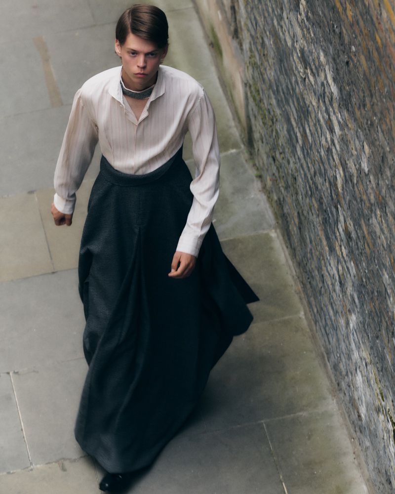 A slender young man walks in a long dark skirt and light shirt on a stone path, with a stone wall behind him, lit by daylight.