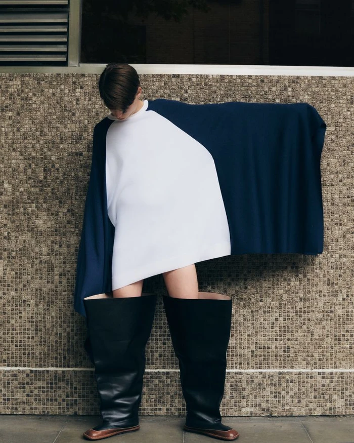 Model Ruben Long posing in an oversized white and navy draped top with giant black leather boots against a tiled wall.