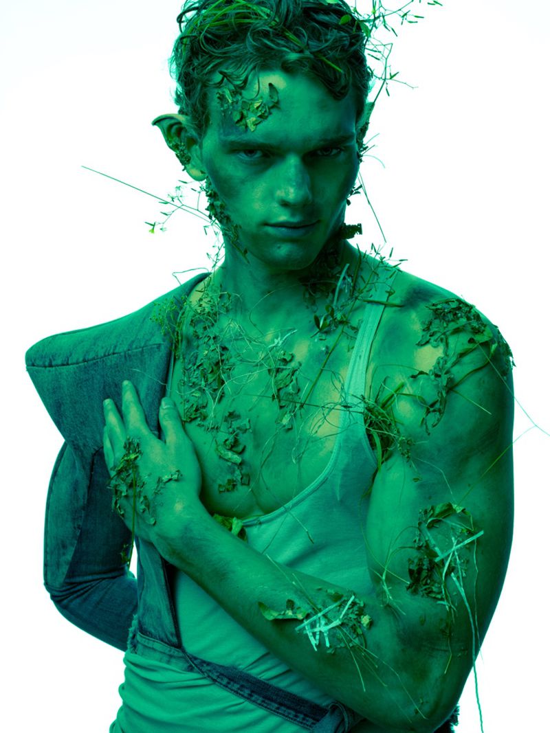 Man with green body paint and grass, wearing a vest, poses against a white backdrop in a surreal fashion or body-art style.