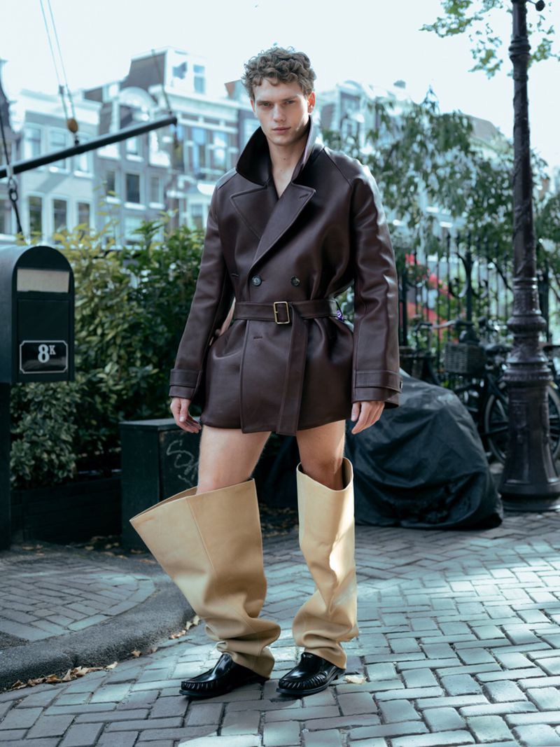 A young adult man models a brown belted jacket and oversize boots on a brick sidewalk in front of townhouses and green trees.