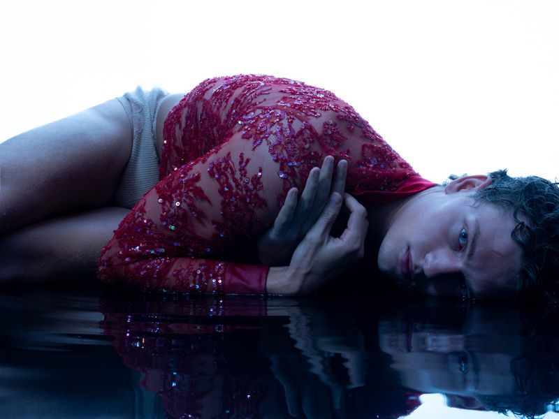 A male model lies prone on a reflective surface wearing a red sequined, sheer bodysuit, with a bright, white background.
