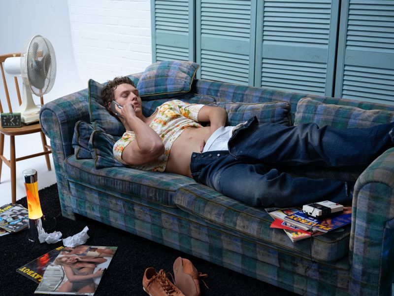 Man lounges on a plaid couch talking on a cell phone, wearing jeans and a floral shirt, with magazines and a fan nearby.