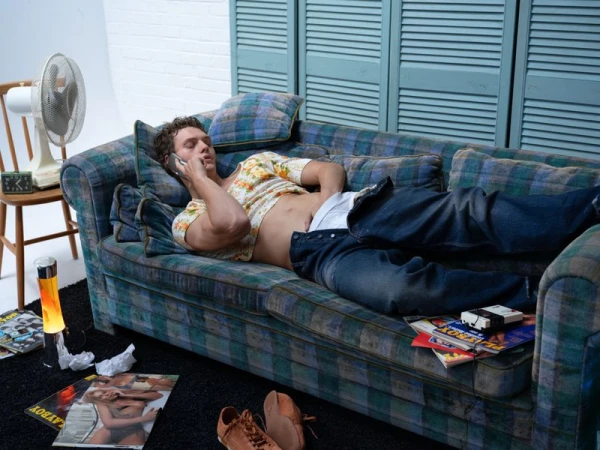 Man lounges on a plaid couch talking on a cell phone, wearing jeans and a floral shirt, with magazines and a fan nearby.