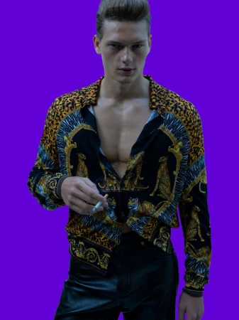 Male fashion model in a leopard-print shirt and leather pants holds a cigarette against a solid purple background in a studio shot.