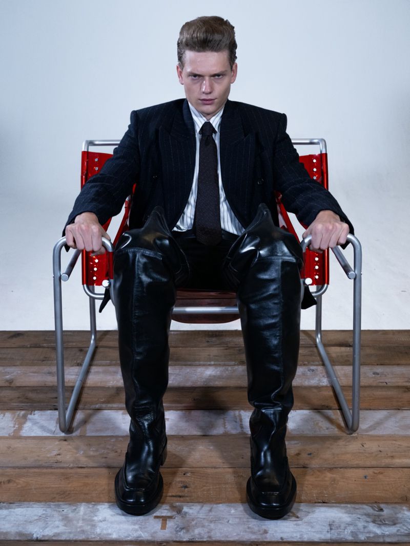 A man wearing tall black boots and a suit sitting in a modern red chair