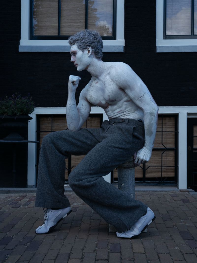 Full-length shot of a shirtless, fair-skinned adult male with stone-like makeup, posed as a statue, seated on a pillar outdoors.