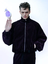 A young male model with curly brown hair wearing a dark purple ribbed zip jacket holding a white and purple pigeon on his hand