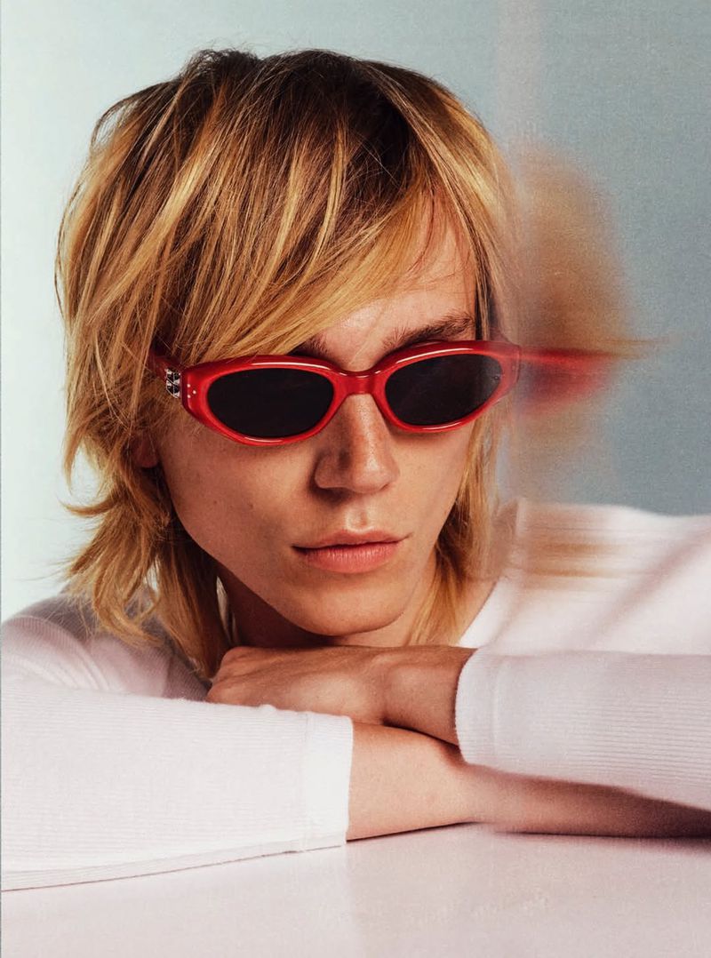 Adult with blonde hair, wearing a white long-sleeved shirt and red sunglasses with dark lenses, resting chin on arms on a white table.