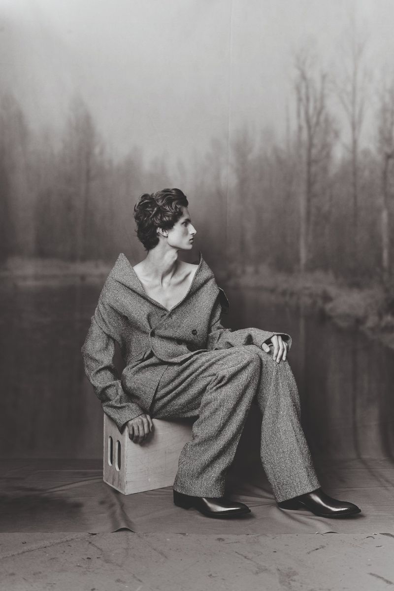 A person in a grey suit poses while sitting on a box in front of a blurry forest background in a studio environment with muted lighting.