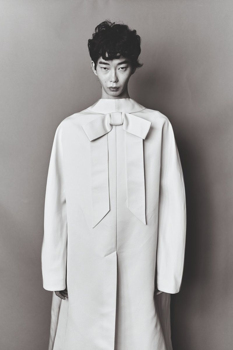 Portrait of a fashion model posing in an elegant, light-colored coat with a large bow detail against a neutral gray backdrop.